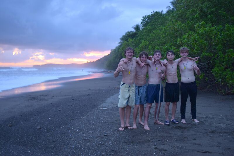 The image shows a group of six young men standing on a black sand beach at dusk. They are shirtless and appear to be enjoying the sunset. The sky is a mix of orange, pink, and purple hues, reflecting on the wet sand. Lush green vegetation covers the hillside in the background, contrasting with the dark beach. The waves are gently rolling in, creating a serene and picturesque scene.